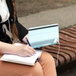 Ręce trzymają tablet i notatnik, na którym robi notatki