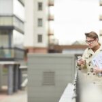 Minimalna talia w górę portret współczesnego młodego biznesmena w płaszczu i trzymającego smartfona, opierając się na poręczy na zewnątrz w miejskim mieście, kopiuj przestrzeń