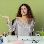 Mylić młoda kobieta całkiem pracownik biurowy siedzi przy biurku z narzędzi biurowych, udając, że trzyma coś na białym tle na oliwkowym tle