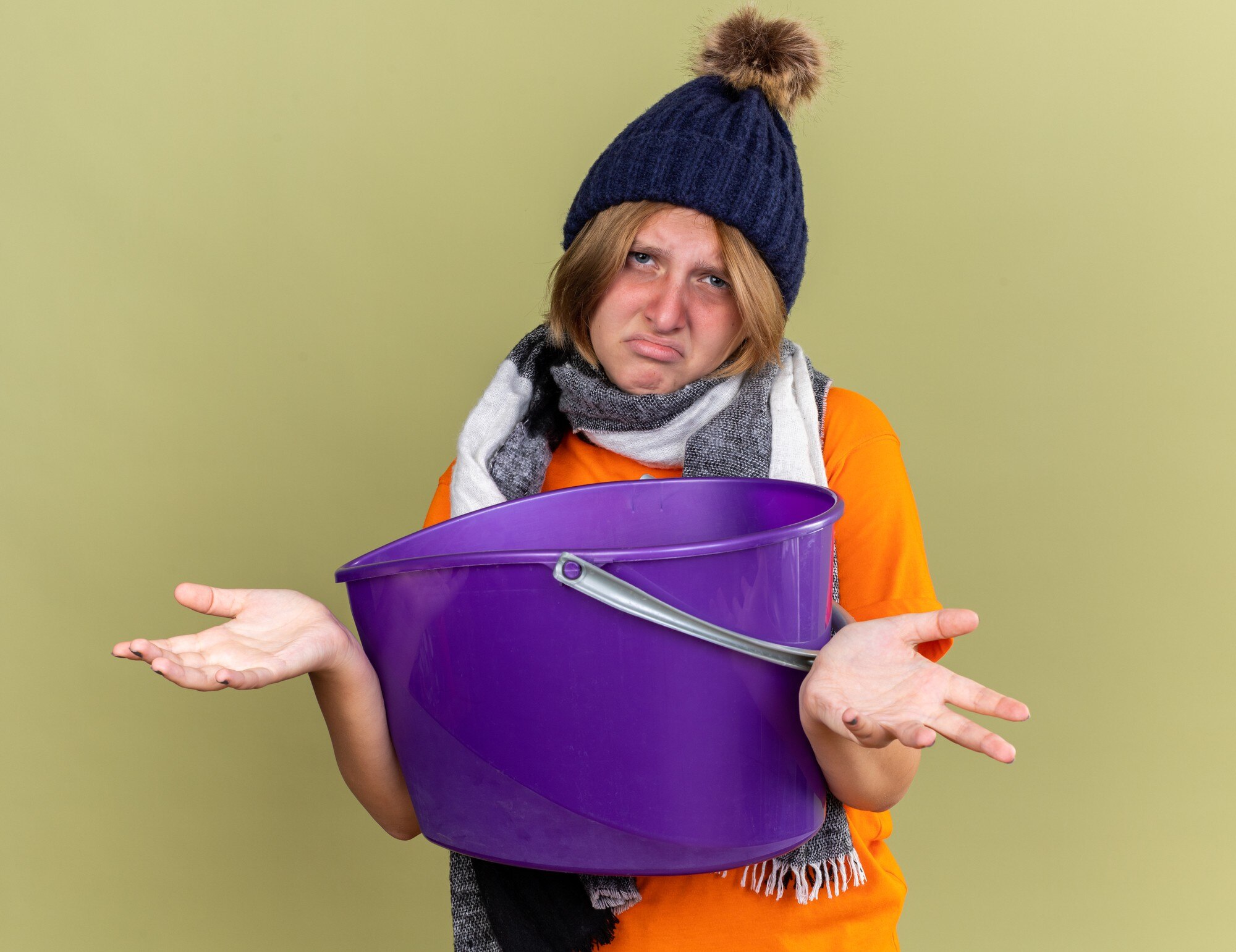 Niezdrowa młoda kobieta w kapeluszu z szalikiem na szyi, źle się czuje, cierpi na mdłości, trzymając basen patrząc zdezorientowana, wzruszając ramionami, stojąc nad zieloną ścianą