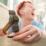 Rudowłosa krótkowłosa kobieta krzyczy z bólu uderzając się w palec młotkiem