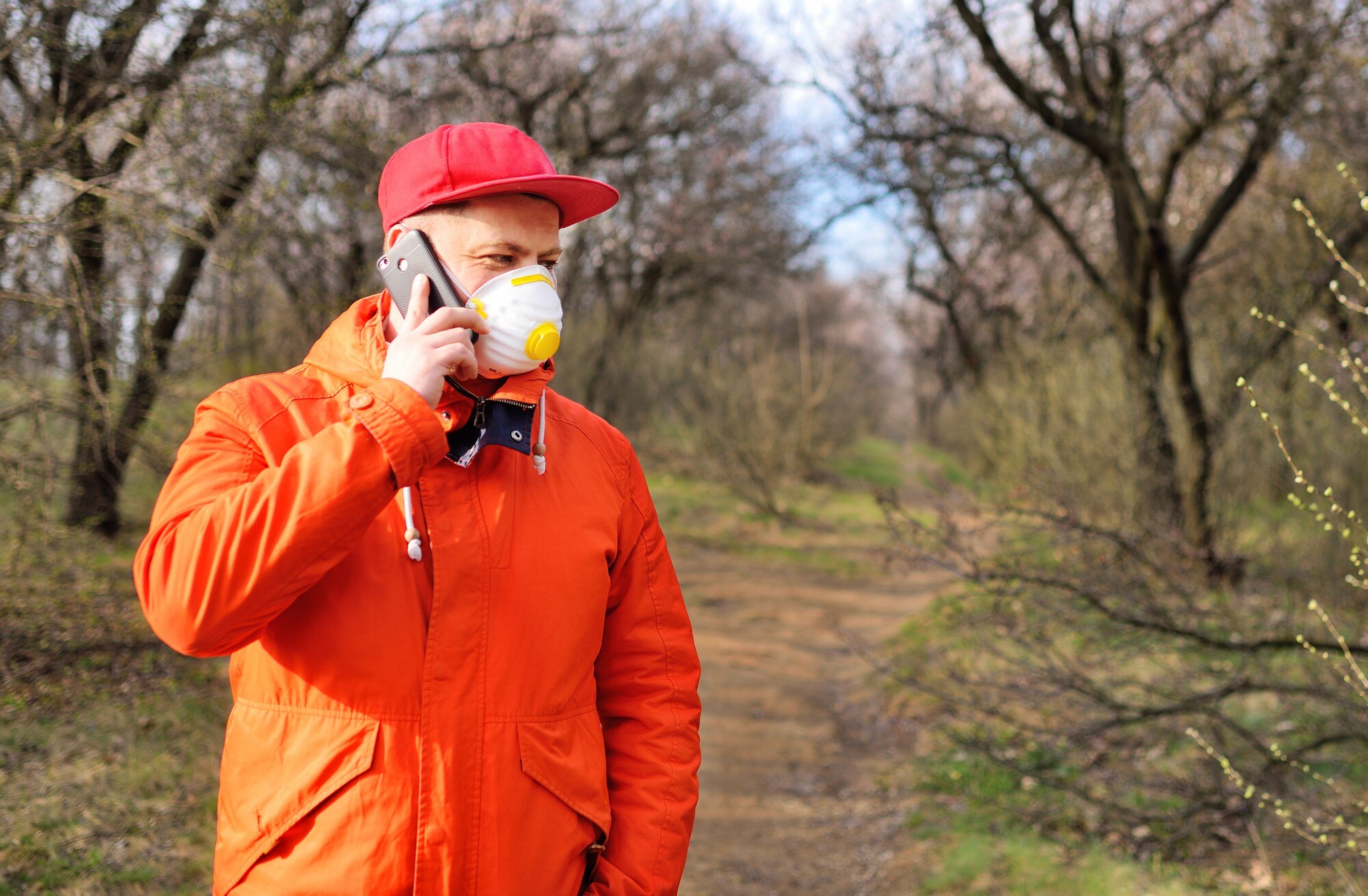 Młody mężczyzna w pomarańczowej kurtce i czerwonej czapce z respiratorem lub maską na twarzy rozmawia przez telefon na tle drzew w parku podczas pandemii koronawirusa Covid-19.
