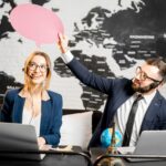 Para travel managerów marząca z kolorowymi bąbelkami nad głową siedząca w biurze agencji na tle mapy świata