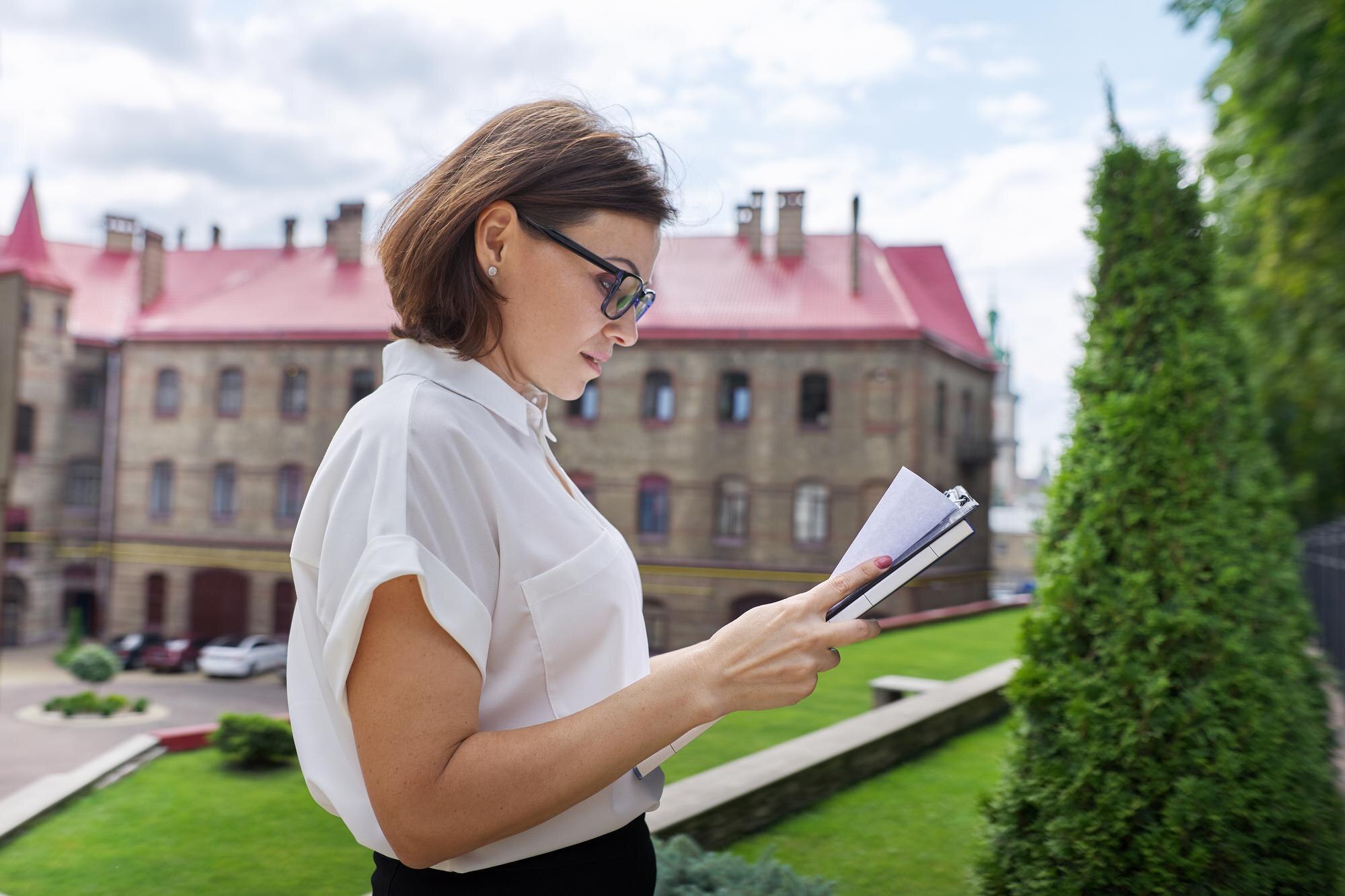 Portret nauczyciela dojrzałej bizneswoman, menedżera z dokumentami dokumentów biznesowych w ręce. Budynek szkoły, tło budynku biurowego, profesjonalna pewna siebie kobieta w okularach biała koszula