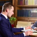 Koncepcja nauki i studiów Mężczyzna w klasycznym garniturze lub profesor ze skoncentrowaną twarzą siedzi w bibliotece w pobliżu stosów książek, półek z książkami na tle, czytanie książki nieostre Mężczyźni uczą się w starej bibliotece