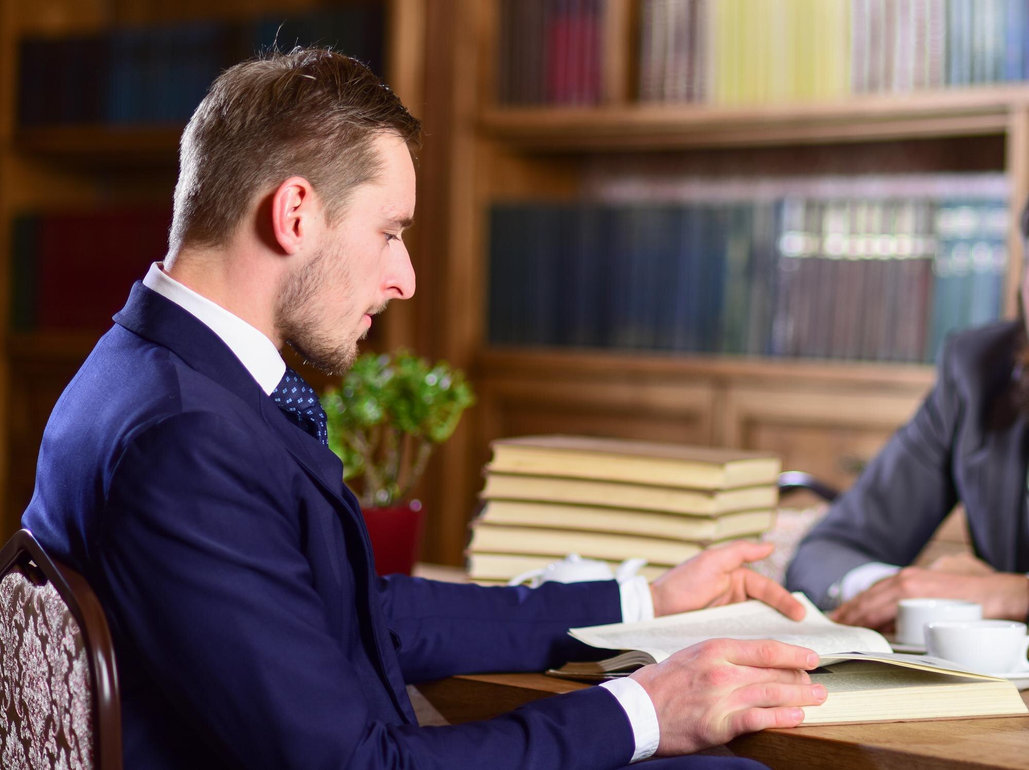 Koncepcja nauki i studiów Mężczyzna w klasycznym garniturze lub profesor ze skoncentrowaną twarzą siedzi w bibliotece w pobliżu stosów książek, półek z książkami na tle, czytanie książki nieostre Mężczyźni uczą się w starej bibliotece