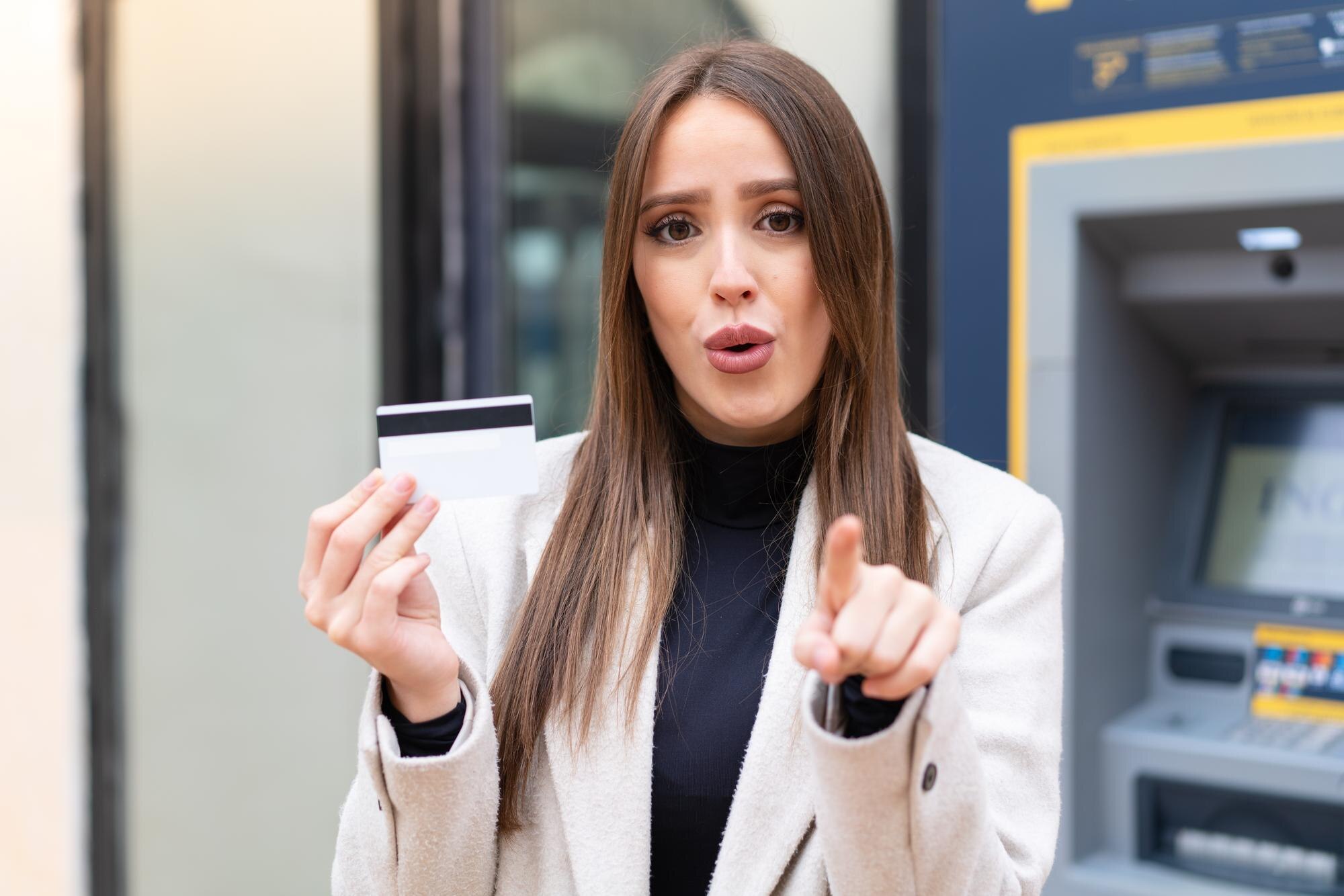Młoda ładna kobieta trzyma kartę kredytową na zewnątrz zaskoczona i wskazująca przód
