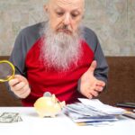 Starszy mężczyzna z szarą brodą analizuje swoje finanse trzymając gotówkę i używając szkła powiększającego