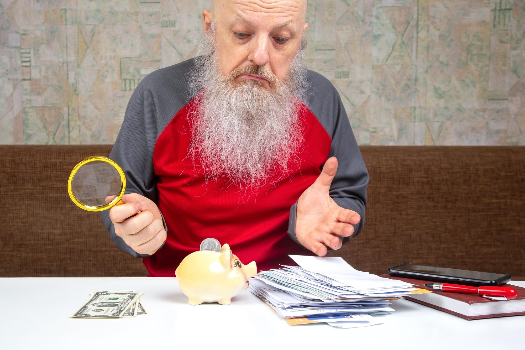 Starszy mężczyzna z szarą brodą analizuje swoje finanse trzymając gotówkę i używając szkła powiększającego