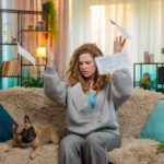 Young woman throws bills on home sofa in frustration while pug dog watches with confused expression