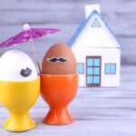 Eggs in egg cups near house on grey wooden background