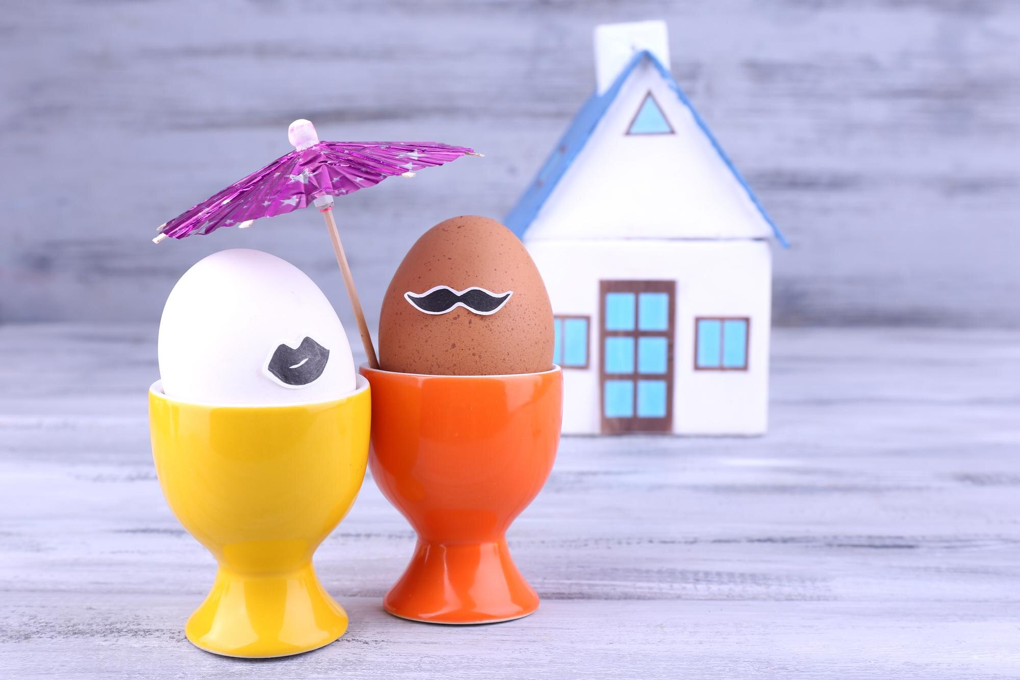 Eggs in egg cups near house on grey wooden background