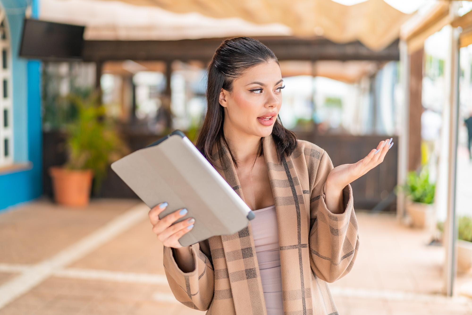 Młoda ładna kobieta trzymająca tablet na świeżym powietrzu ze zdziwionym wyrazem twarzy