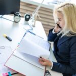Młoda dziewczyna siedzi przy biurku w biurze, rozmawia przez telefon i patrząc na dokumenty.