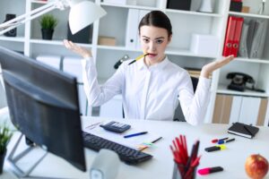 Młoda dziewczyna w biurze siedzi przy stole, trzymając w ustach długopis i rozkładając ręce na boki.